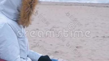穿着白色<strong>羽绒服</strong>的女人坐在海滩上的长凳上戴上手套。 海冬，冻女人..